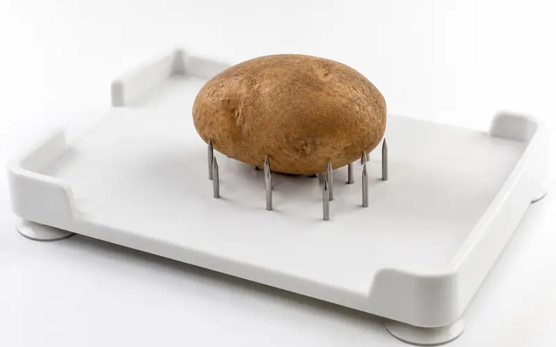 Illustration of a white food preparation board with spikes holding a potato. Shows the type of product, not the exact brand or model.