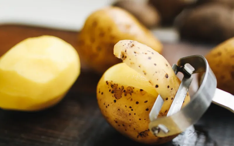 Photo of a potato being peeled with a metal peeler on a wooden board. A general kitchen photo, not this product.