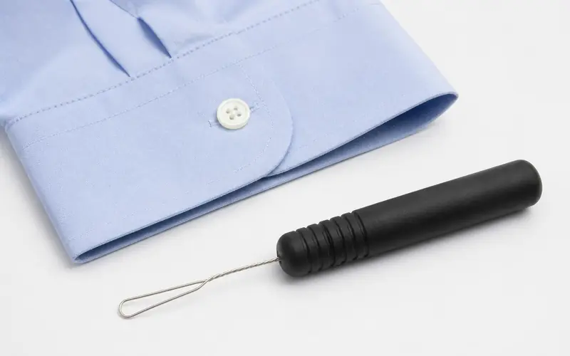 Illustration of a button hook with a thick black grip beside a shirt cuff. Shows the type of product, not the exact brand or model.